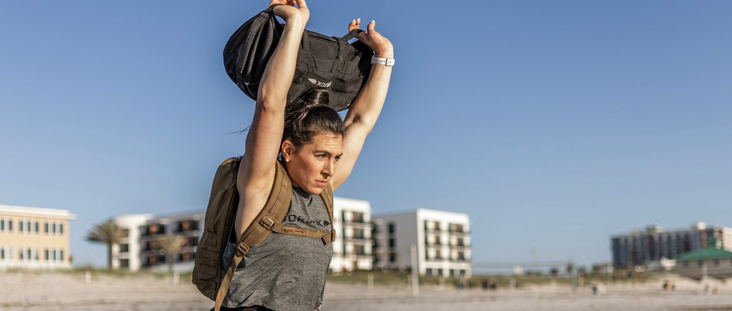 Woman rucking outdoors with GORUCK gear, lifting sandbag overhead on sunny beach