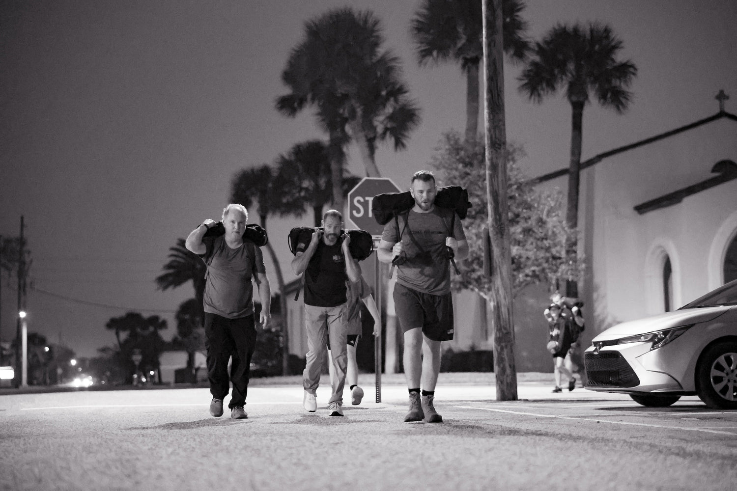 Three men rucking with sandbags on a city street at night, palm trees in background, GORUCK gear