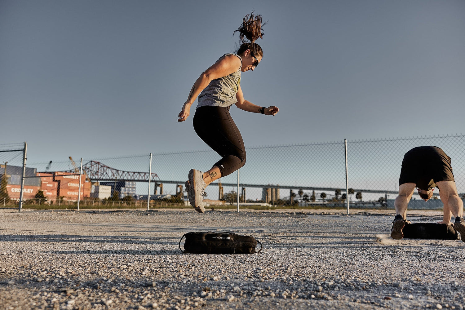 Woman jumping over GORUCK sandbag outdoors during rucking workout with shipping containers in background.