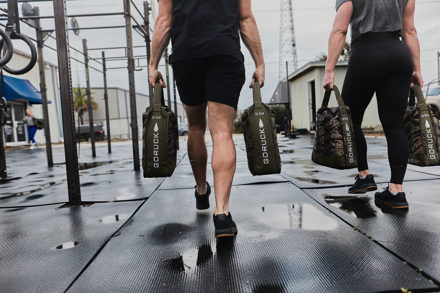 Two athletes carrying GORUCK sandbags at an outdoor gym, showcasing rugged rucking gear.