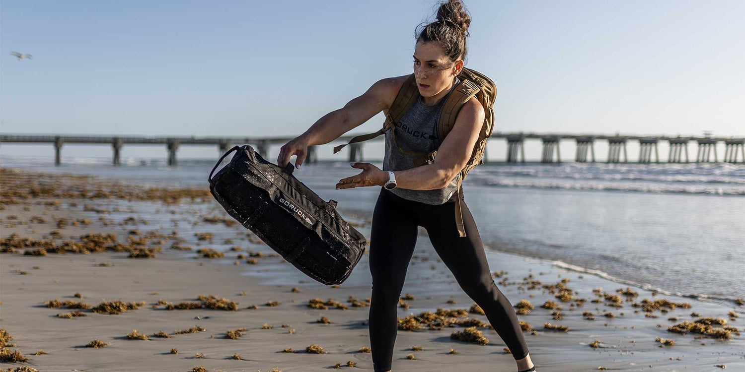 Woman training on beach with GORUCK sandbag and backpack, pier and ocean in background