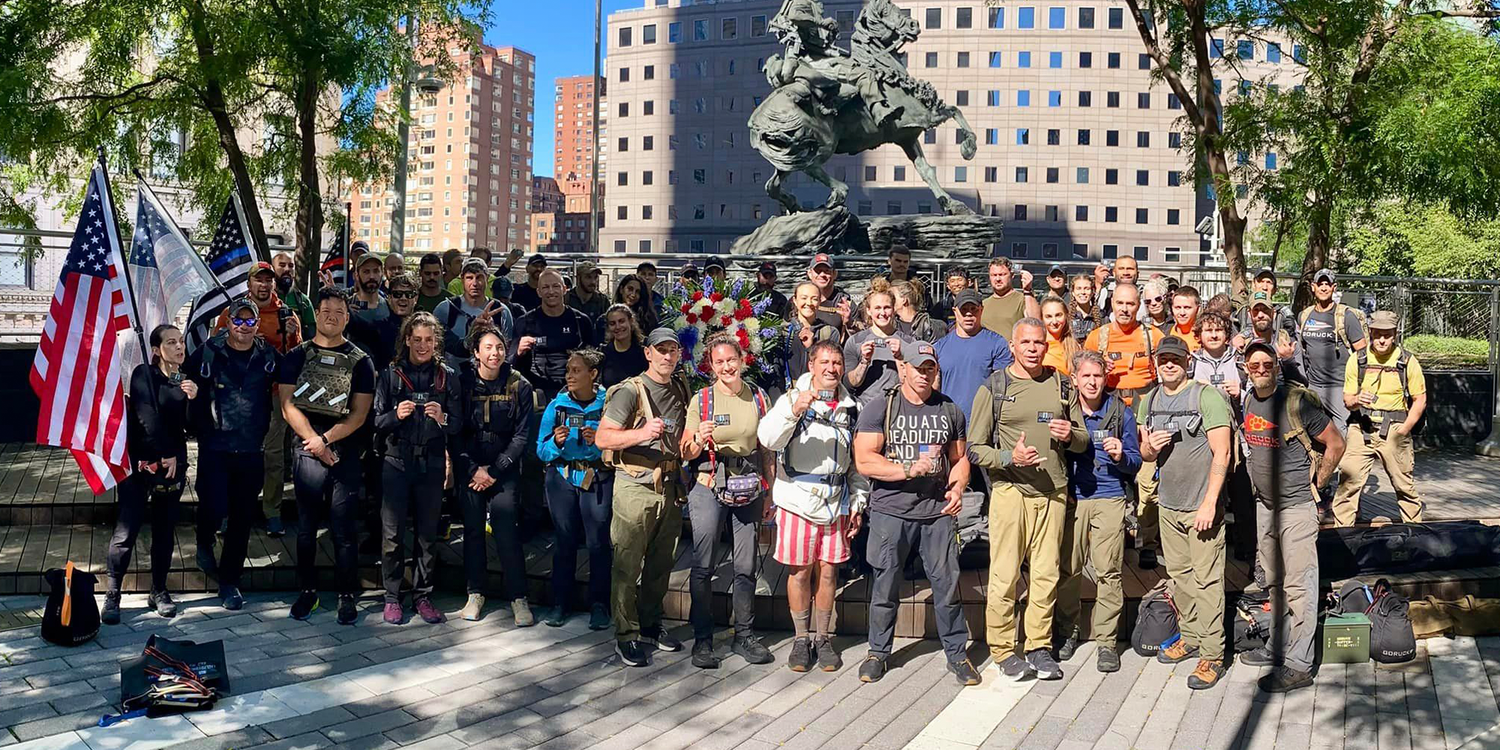 GORUCK group event participants with American flags, rucking gear, at outdoor memorial plaza