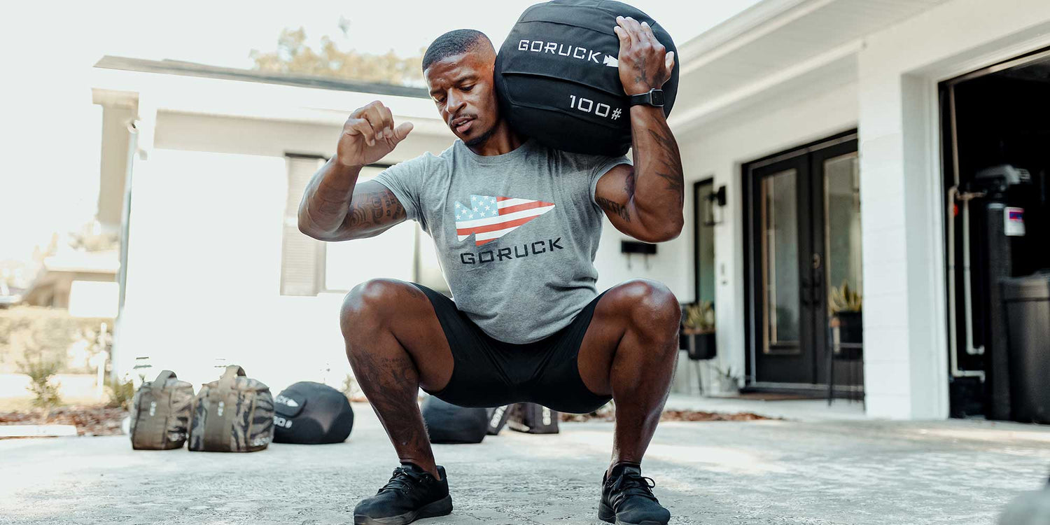 Fit man doing a weighted squat holding a 100-pound GORUCK sandbag outdoors