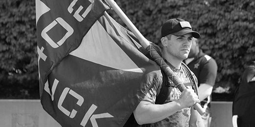 Black and white photo of a man wearing a cap holding a large flag outdoors with trees in the background