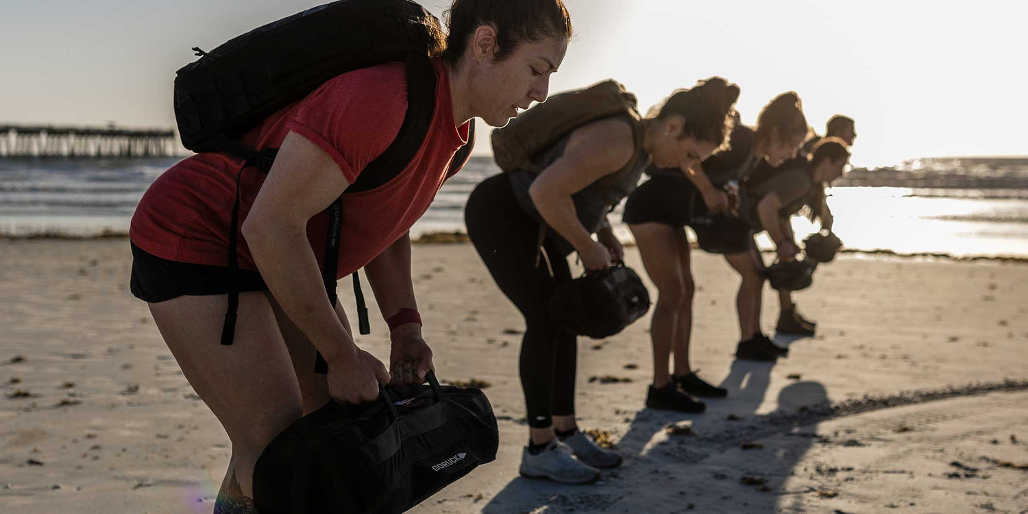 Group of women exercising on beach lifting weighted bags during sunset fitness training