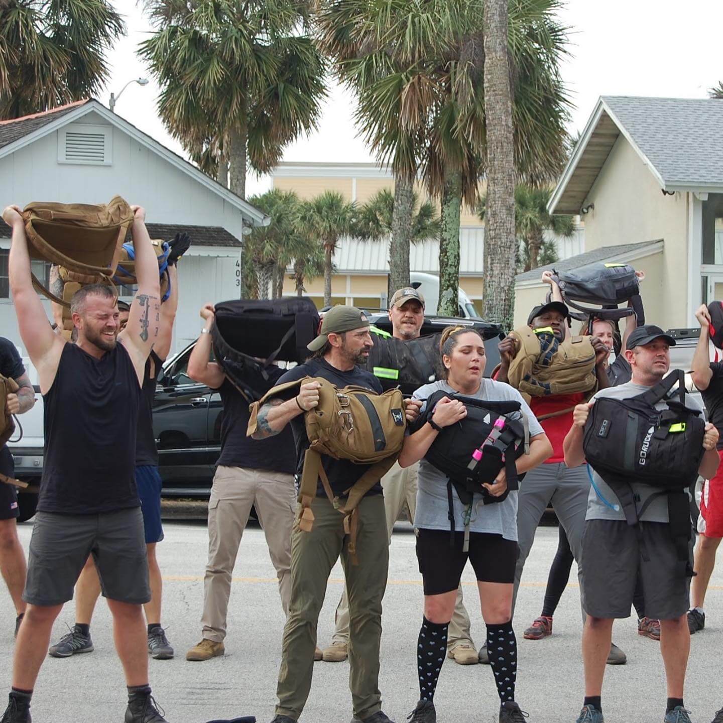 Group rucking workout outdoors with GORUCK gear, participants lifting backpacks, palm trees