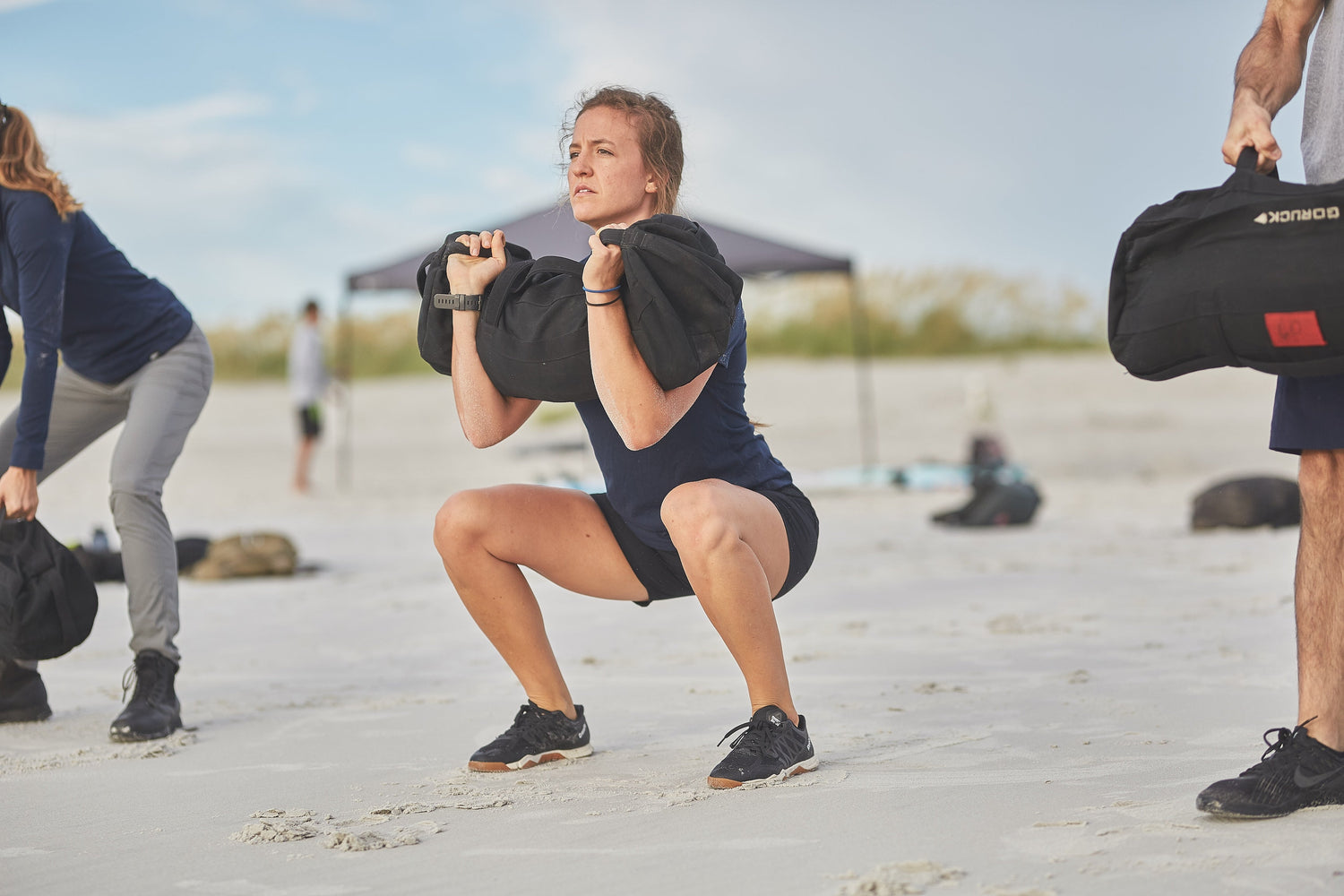 Woman doing sandbag squat on beach during GORUCK outdoor workout event