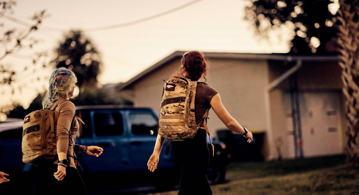 Two women walking outdoors at dusk wearing GORUCK tactical backpacks with a blue truck and house in the background