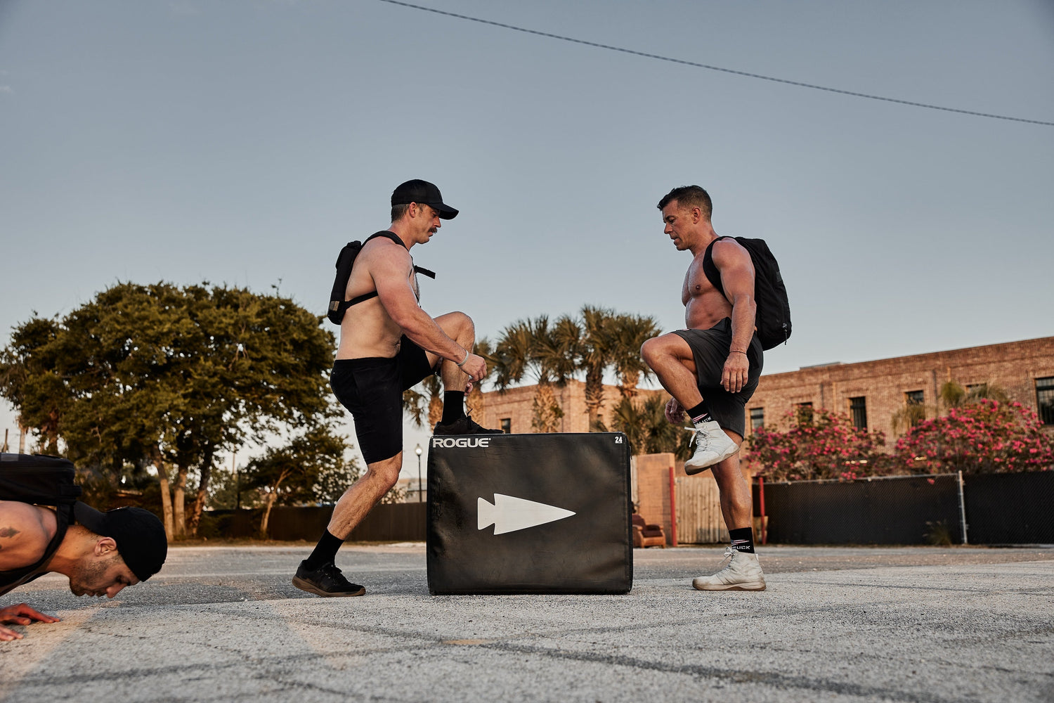 Two men wearing GORUCK rucksacks exercise outdoors with a Rogue plyo box, another man doing push-ups.
