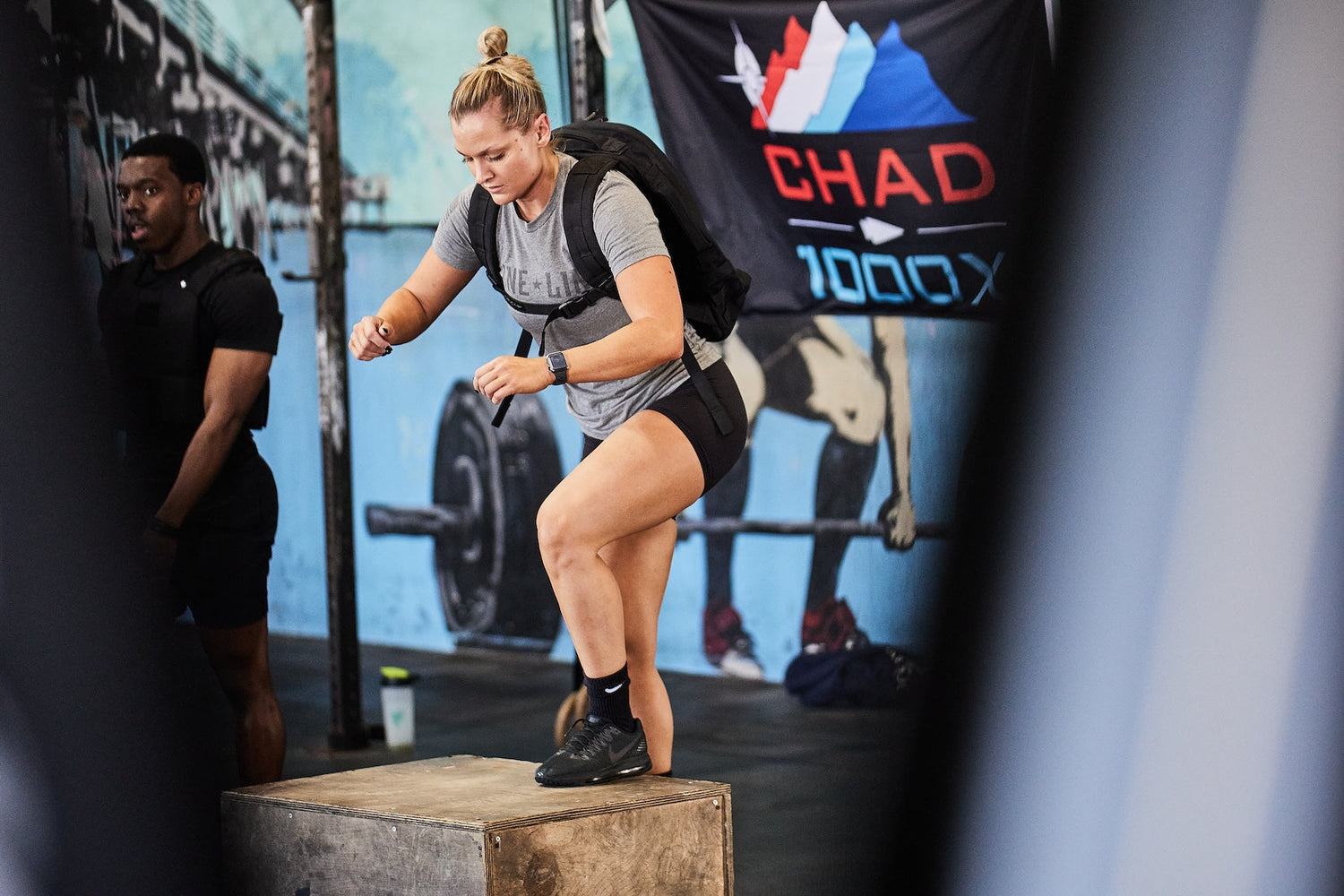 Athlete with rucksack doing box step-ups at gym, GORUCK CHAD 1000X event banner in background