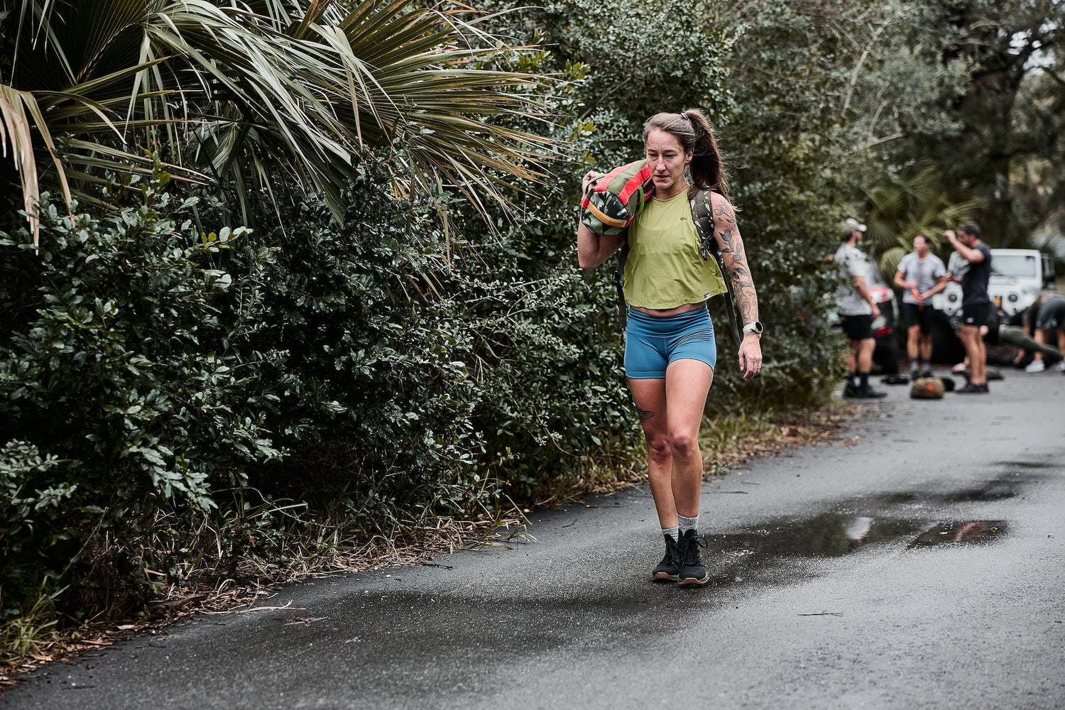 Woman rucking with sandbag on rural road, GORUCK outdoor training event, fitness gear.