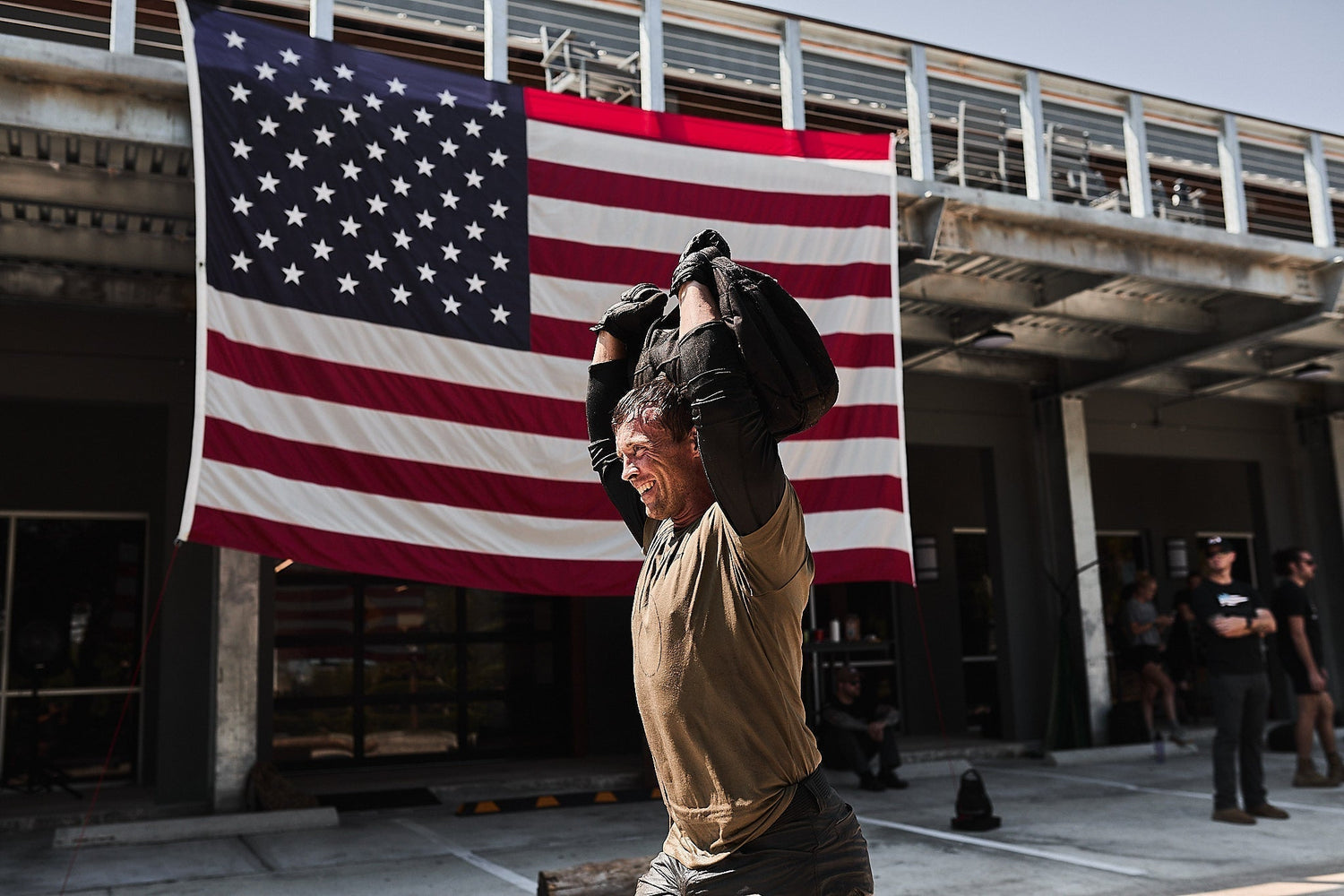 Man lifting weighted rucksack outdoors, GORUCK event, large American flag in background.