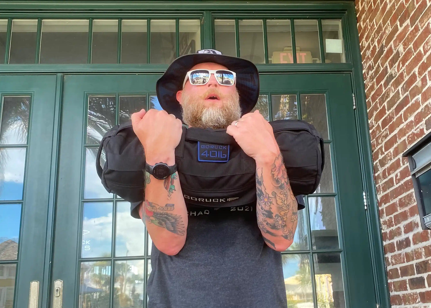 Man with tattoos using GORUCK sandbag for rucking workout outdoors, wearing sunglasses and hat