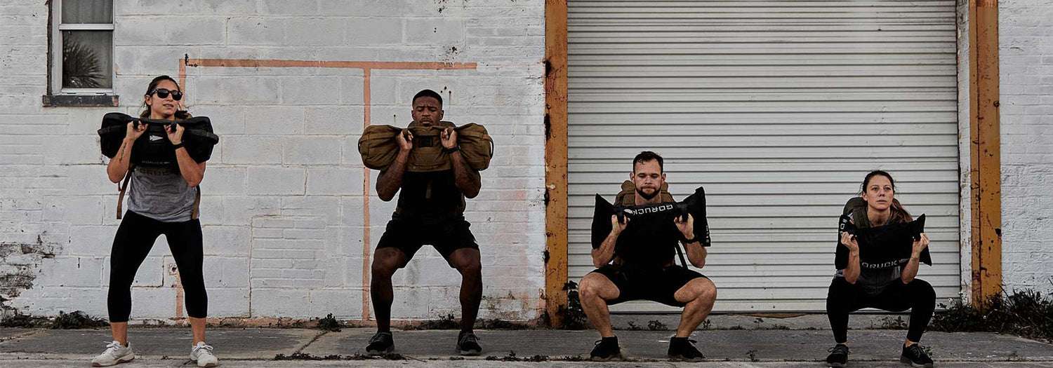 Four people exercising outdoors doing weighted squats with sandbags against a brick wall and garage door