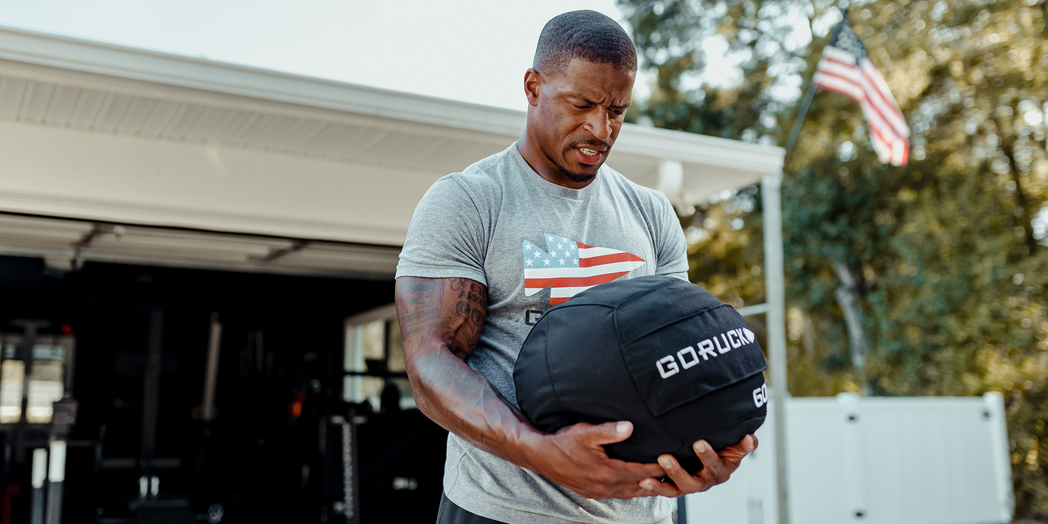 Man lifting GORUCK sandbag outside garage gym, wearing American flag t-shirt, US flag in background