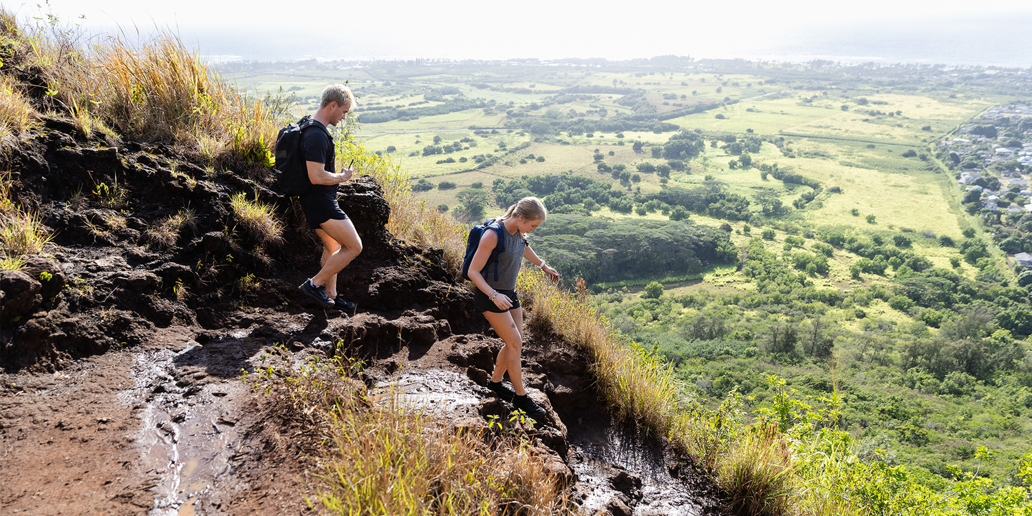 Two hikers descending steep rocky trail wearing backpacks and hiking shoes overlooking green valley