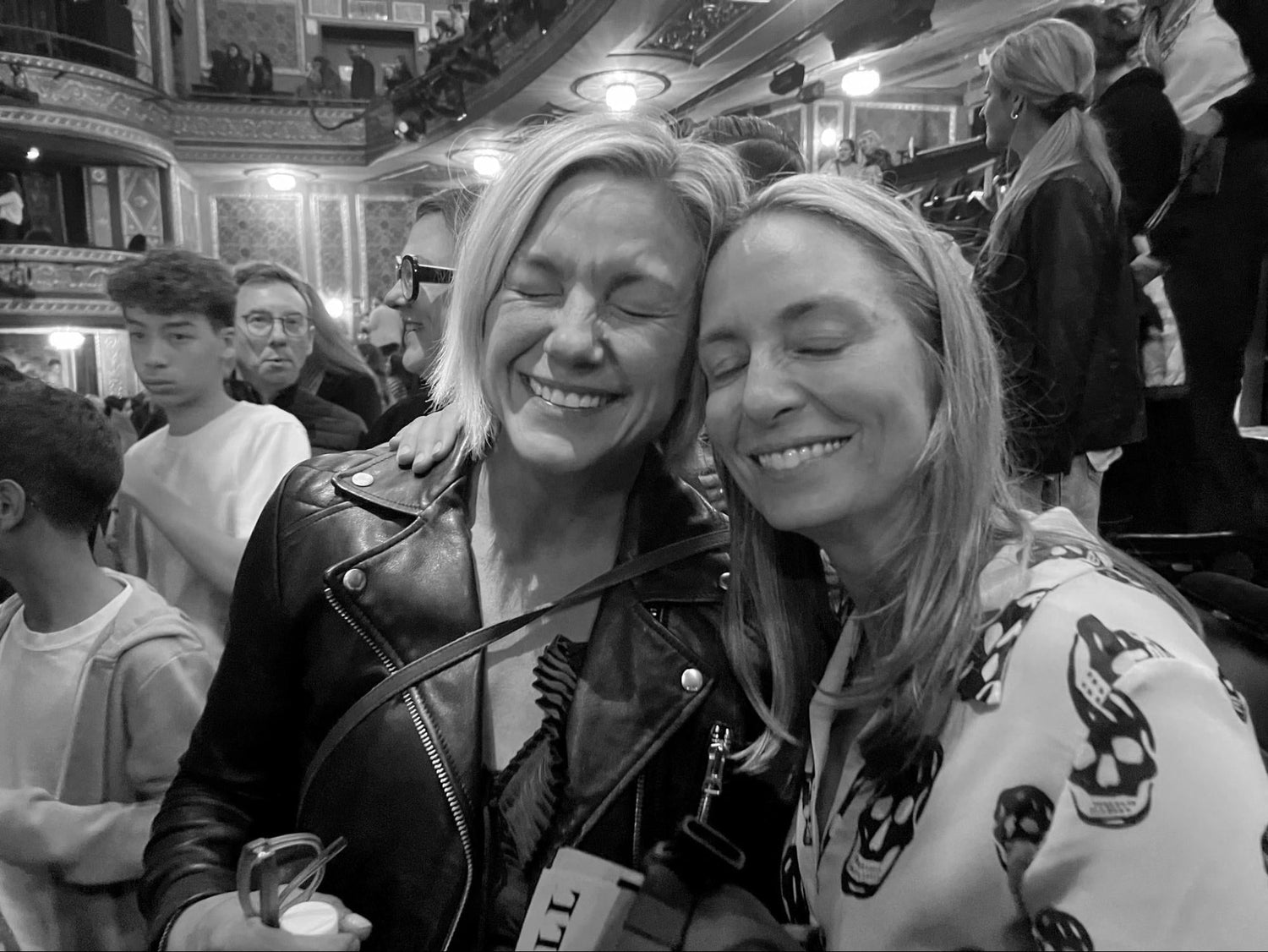 Two smiling women with closed eyes in a crowded ornate theater interior, one wearing a leather jacket, the other a skull-print hoodie