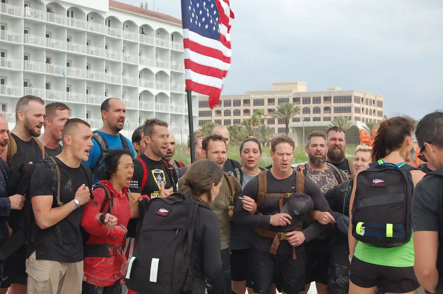 Group of GORUCK participants with backpacks and American flag on beach, rucking event, community focus