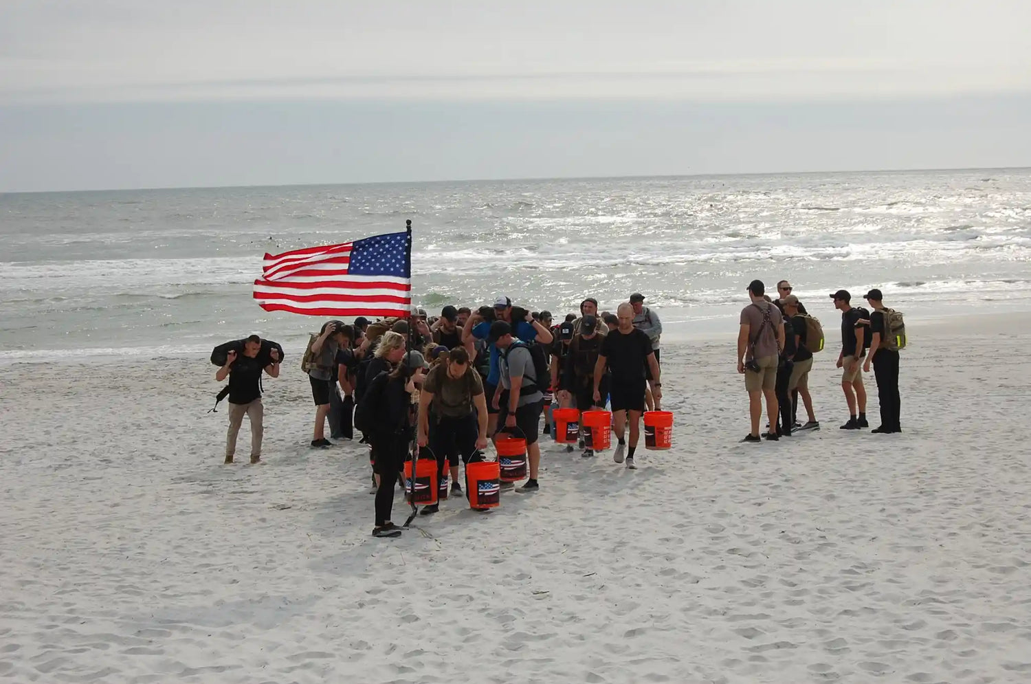 Group of people on a sandy beach carrying weighted buckets and an American flag, engaged in rucking challenge