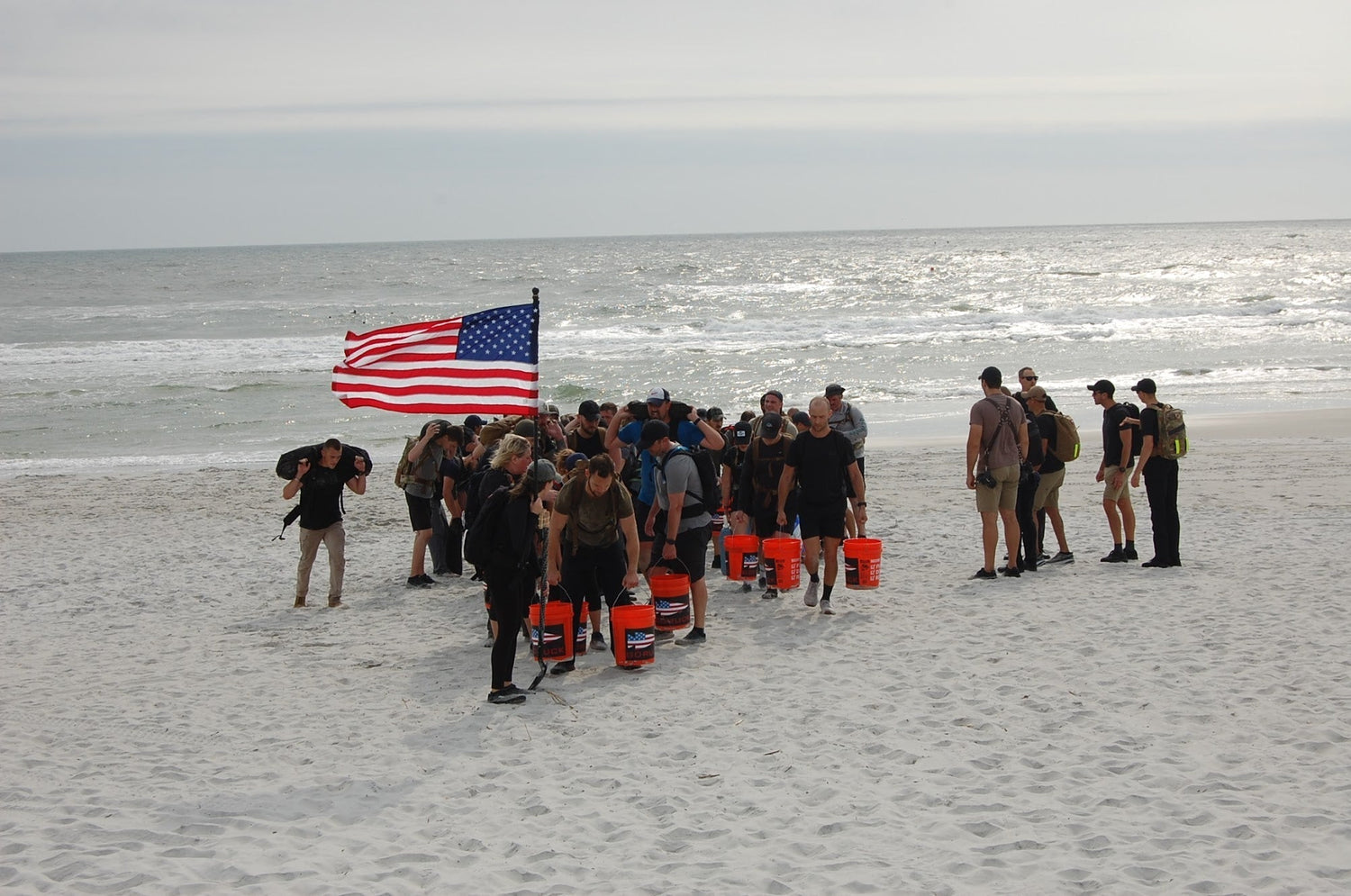 GORUCK team rucking on a beach with backpacks, orange buckets, and an American flag.