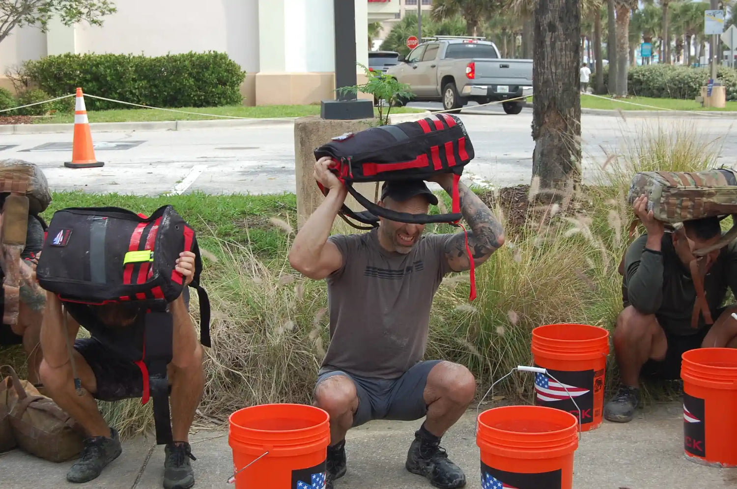 Group of people squatting outdoors with GORUCK rucksacks over their heads during a fitness event