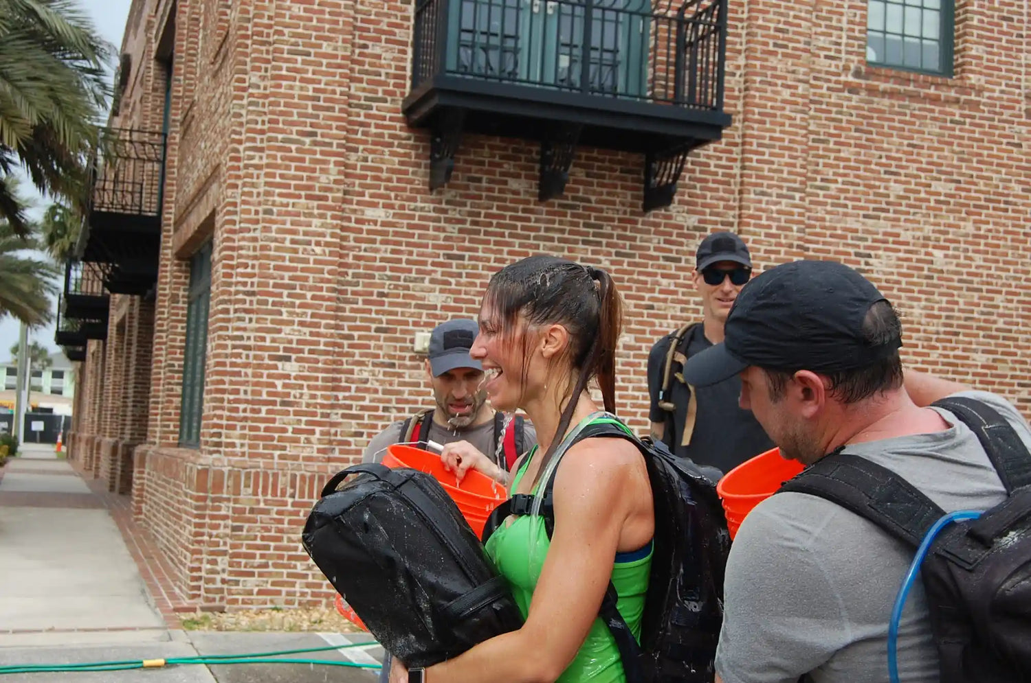 Rucking team in wet GORUCK gear training outdoors near a brick building with orange buckets