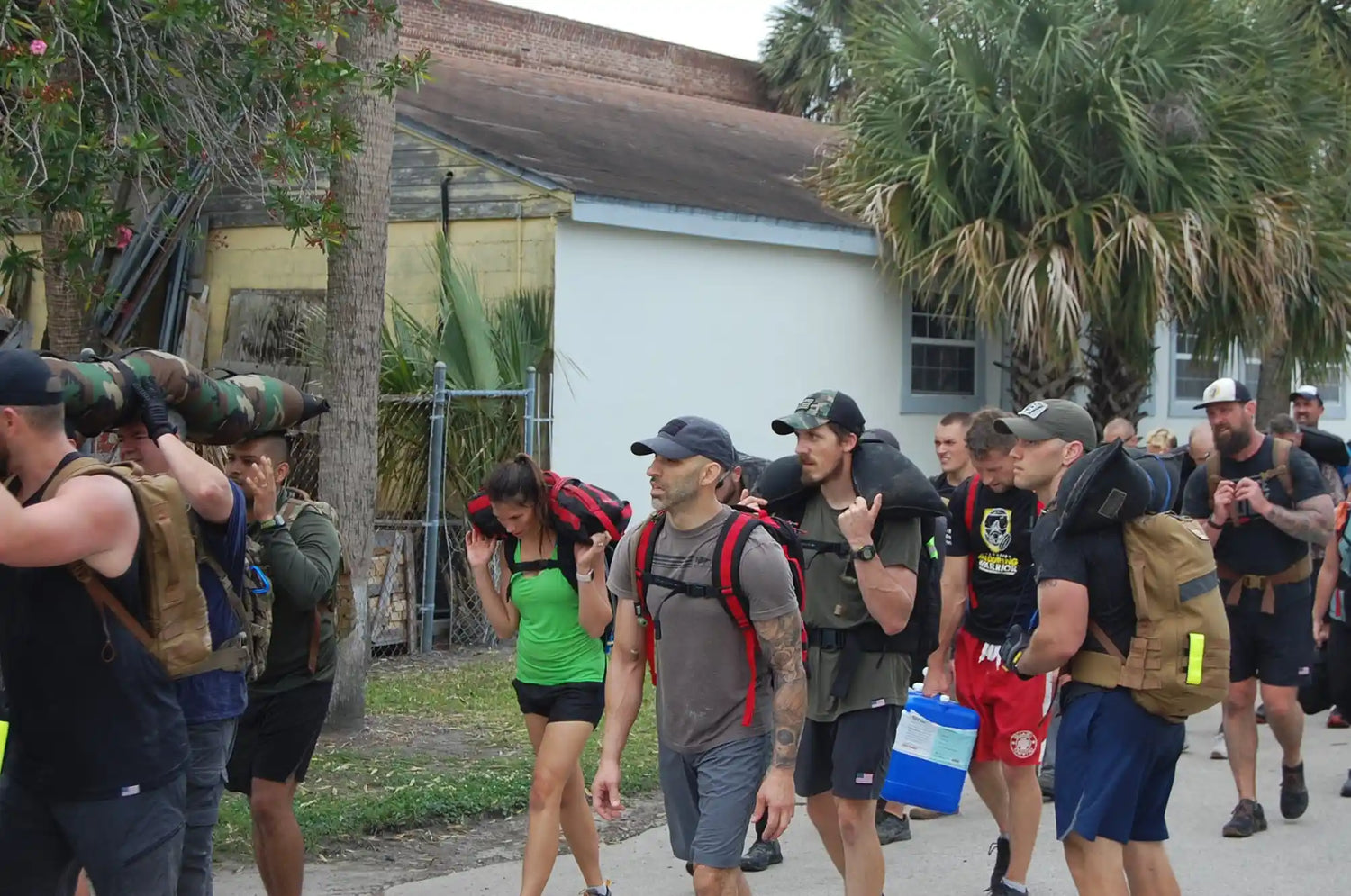 Group of people carrying heavy sandbags and backpacks outdoors near palm trees and buildings