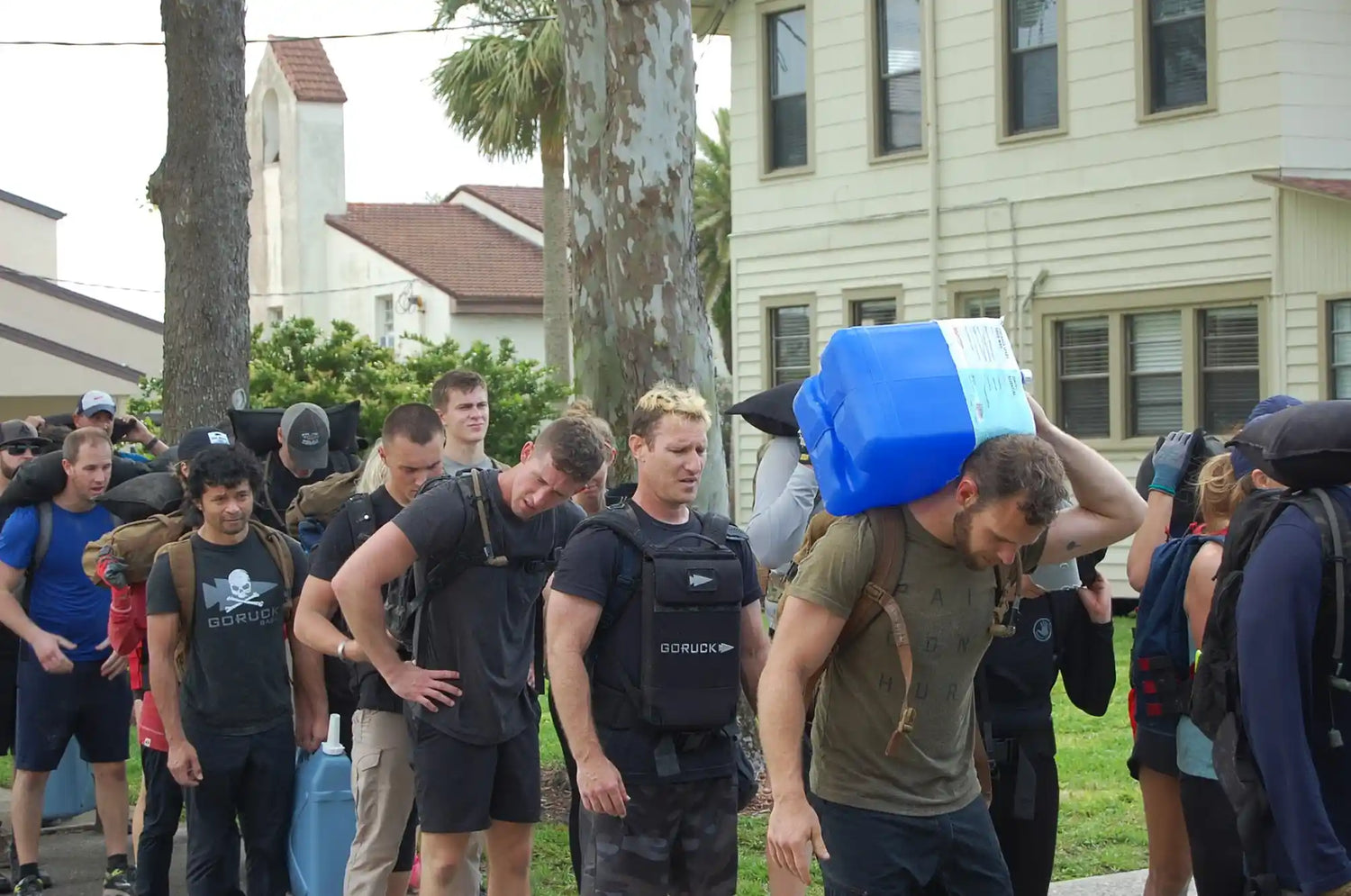 GORUCK participants in outdoor rucking event carrying heavy gear and water jugs in a line