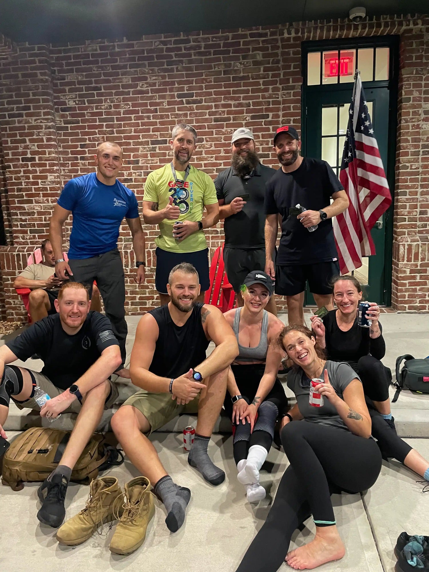Group of nine smiling friends in casual sportswear sitting and standing outside brick building with American flag