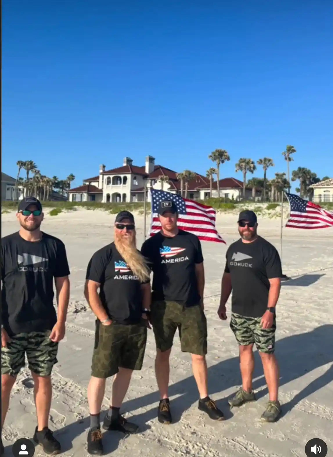 Four men in GORUCK shirts and camo shorts stand on a beach with American flags behind them.