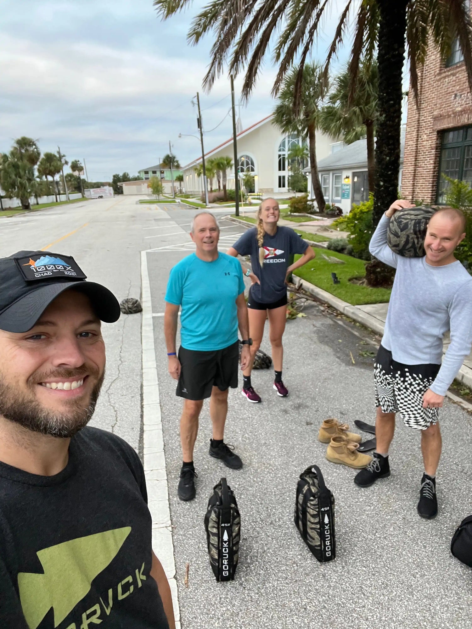 Four people outdoors on a street with palm trees, two holding GORUCK weighted bags for fitness training