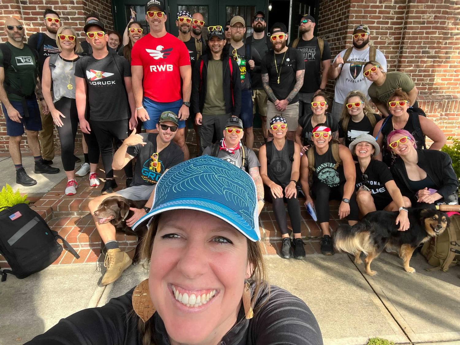 Smiling woman in blue hat taking selfie with group wearing matching yellow sunglasses outside brick building