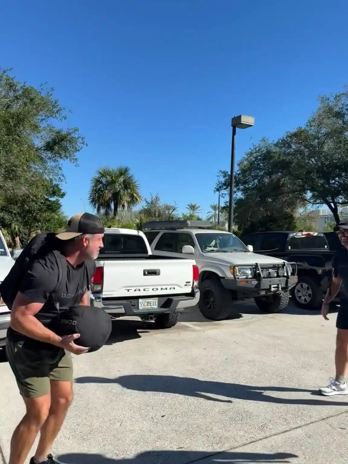 Man with GORUCK gear lifting sandbag outdoors in parking lot with palm trees