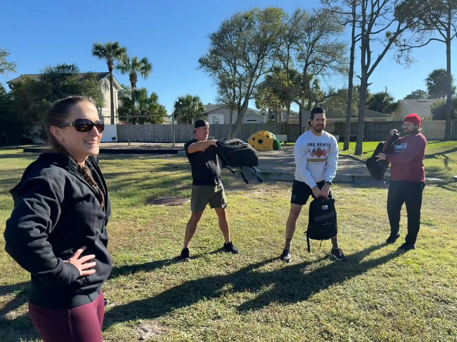 Group training outdoors in sunny park with participants holding and swinging GORUCK backpacks