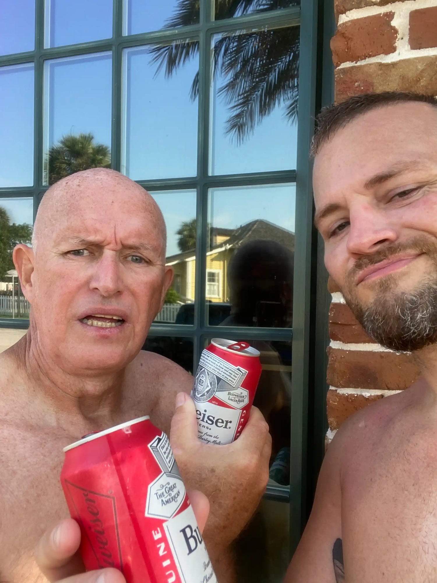 Two shirtless men holding Budweiser beer cans outdoors near window and brick wall under blue sky