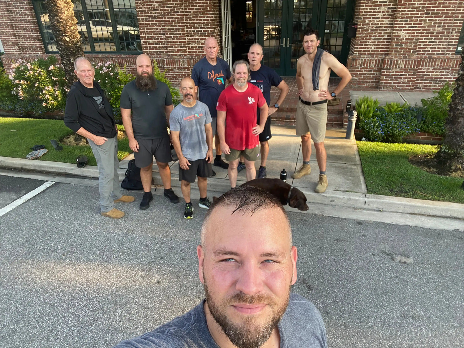 Group of men in casual workout clothes standing outside brick building with greenery and dog on sidewalk