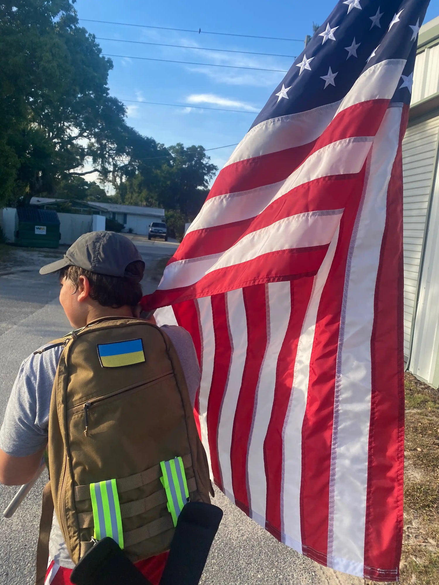 Person wearing backpack with Ukraine patch holding large American flag outdoors on sunny day