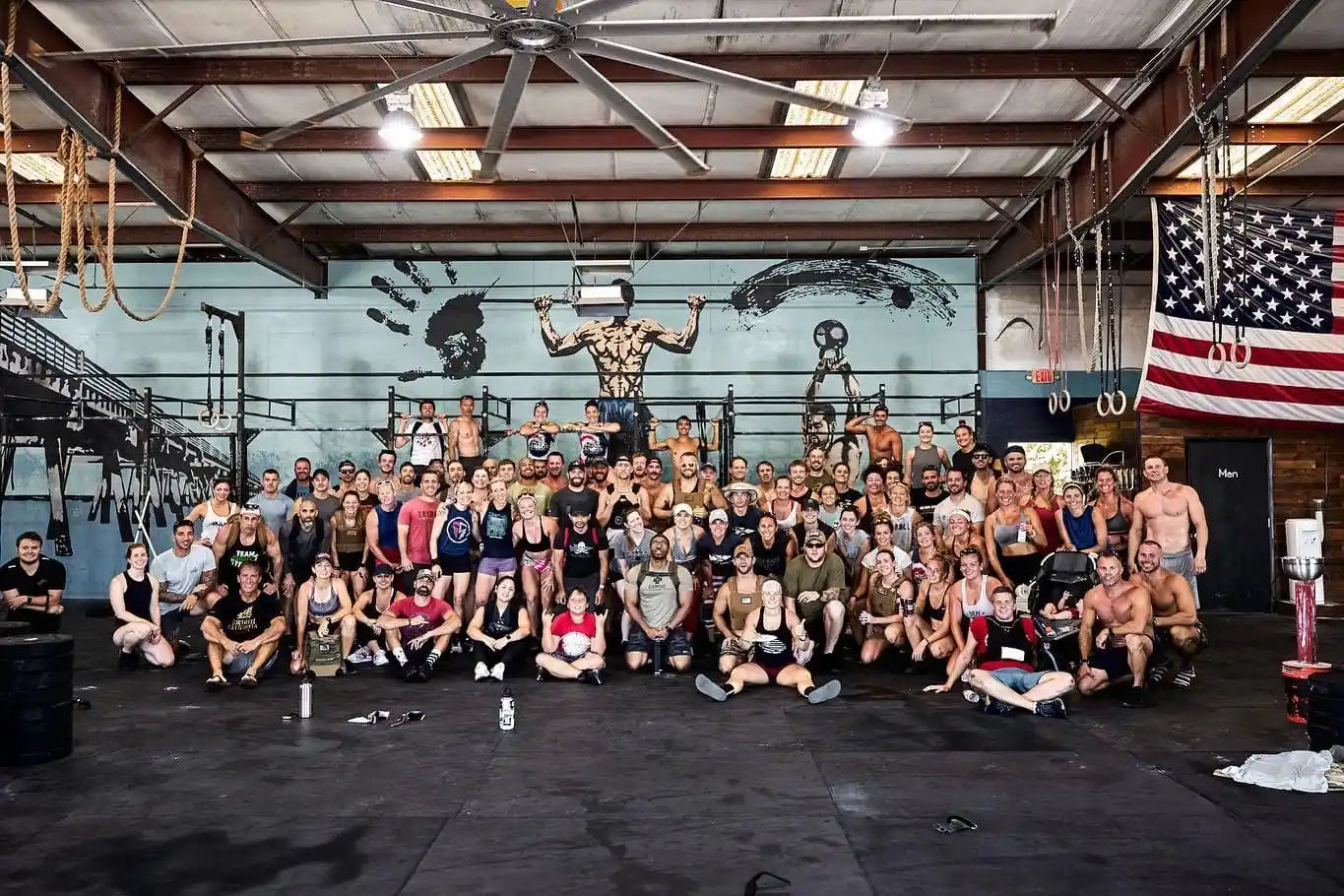 Large group of diverse men and women in athletic wear posing inside a CrossFit gym with industrial ceiling and American flag