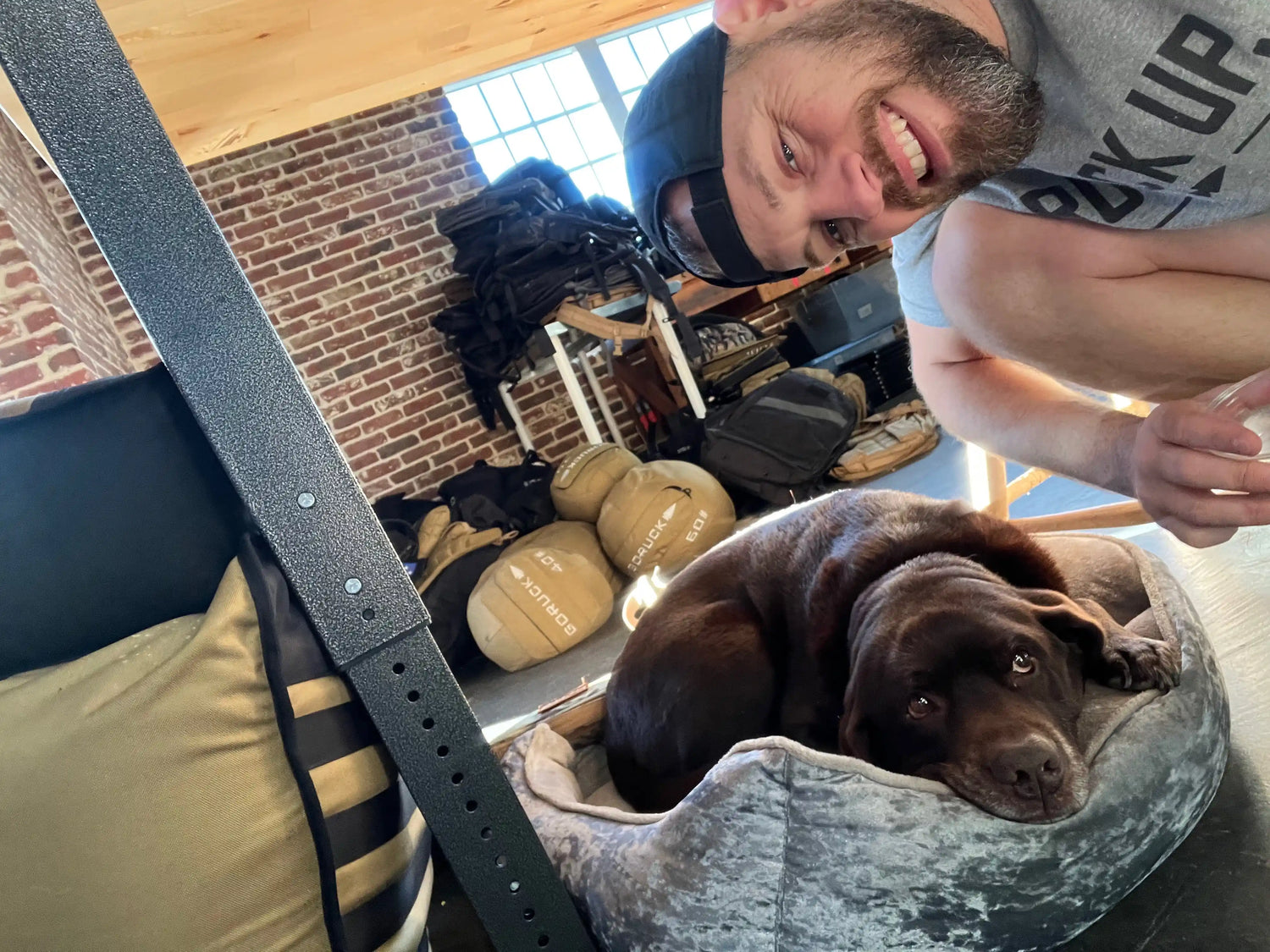 Smiling man in GORUCK shirt with backwards cap next to brown dog resting in a plush bed inside a brick-walled gym