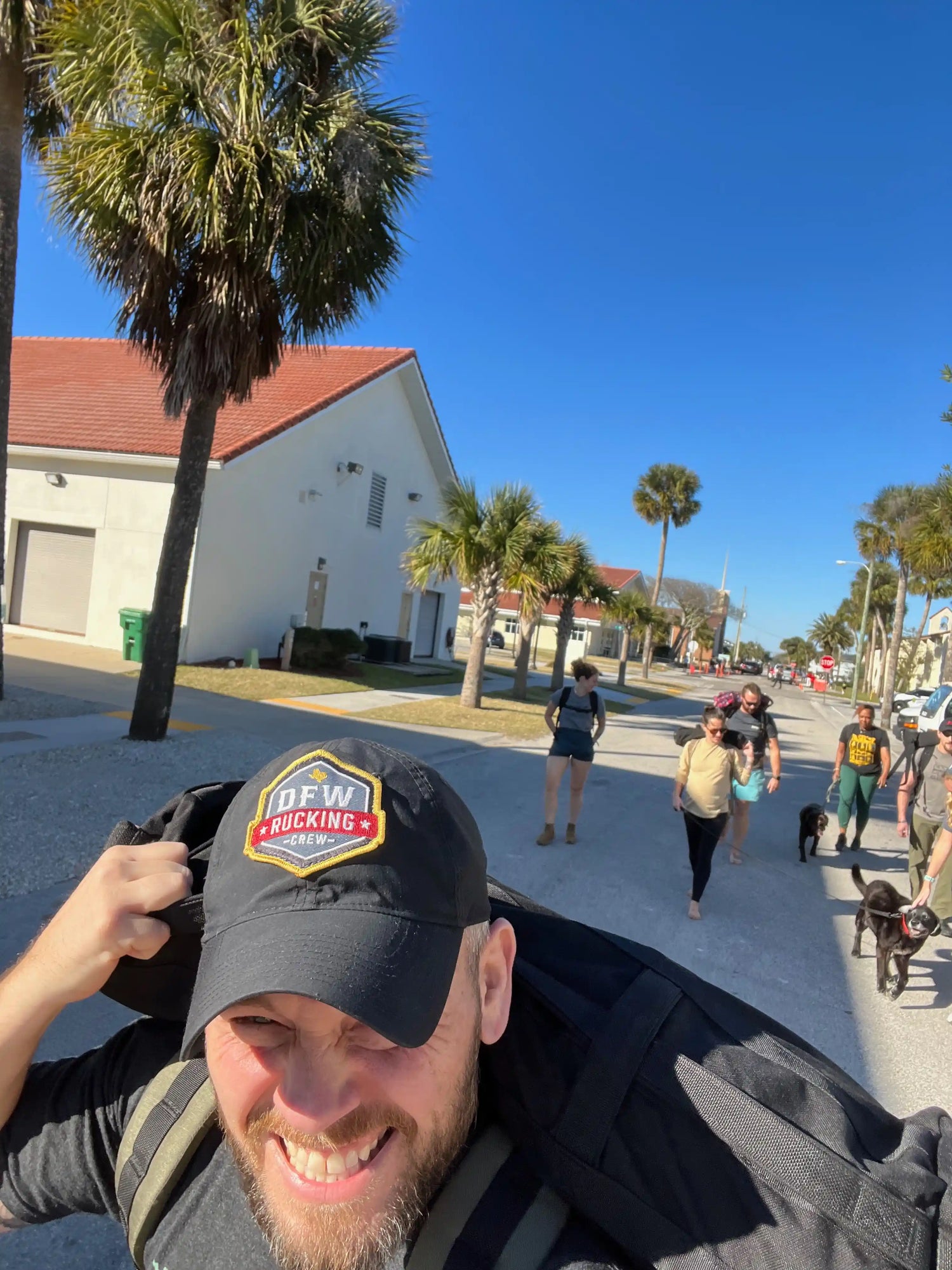 Man wearing DFW rucking crew cap carrying heavy ruck near palm trees with group rucking down sunny street
