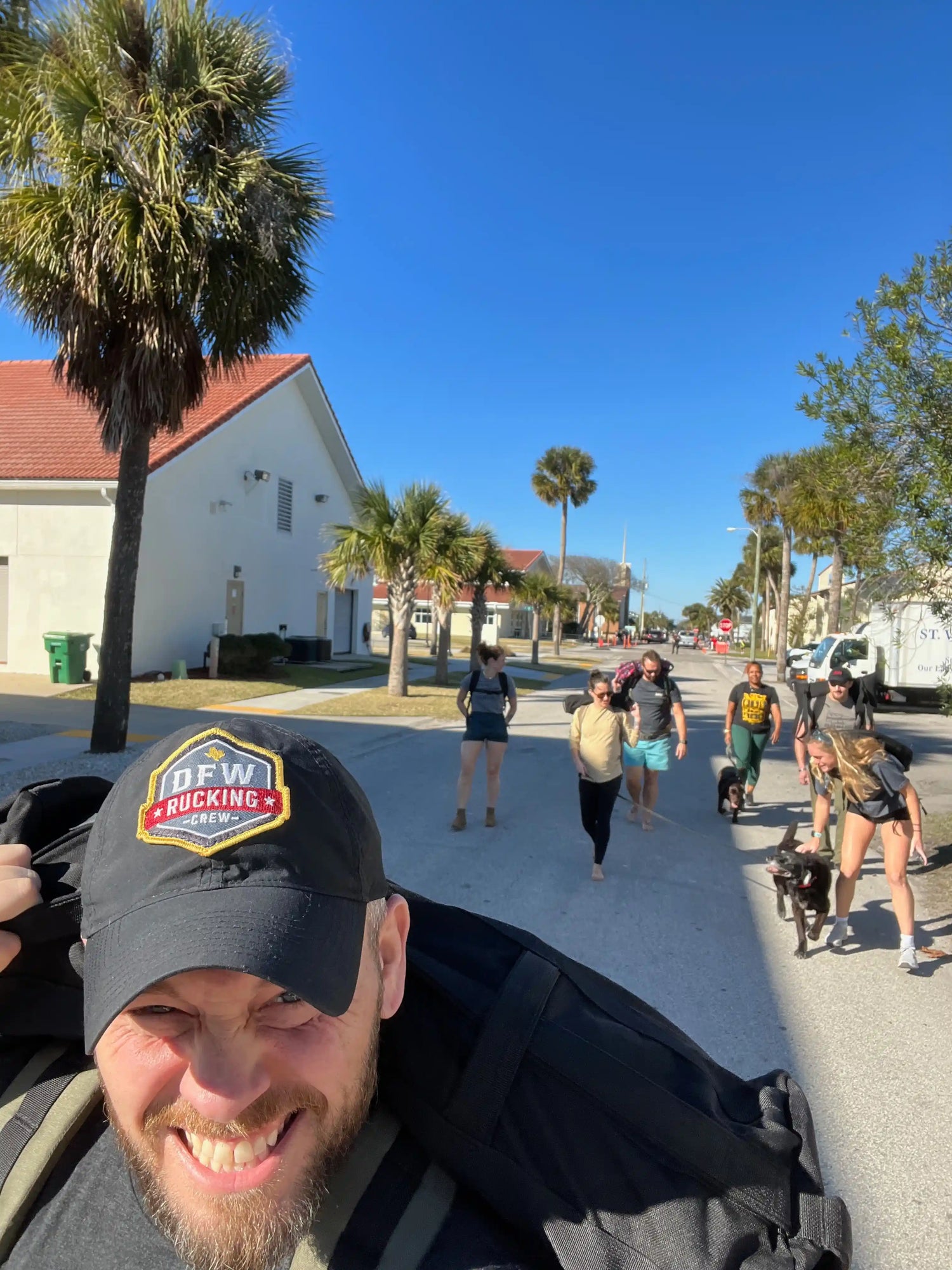 Smiling man in black cap with backpack leading group walking on sunny street with palm trees