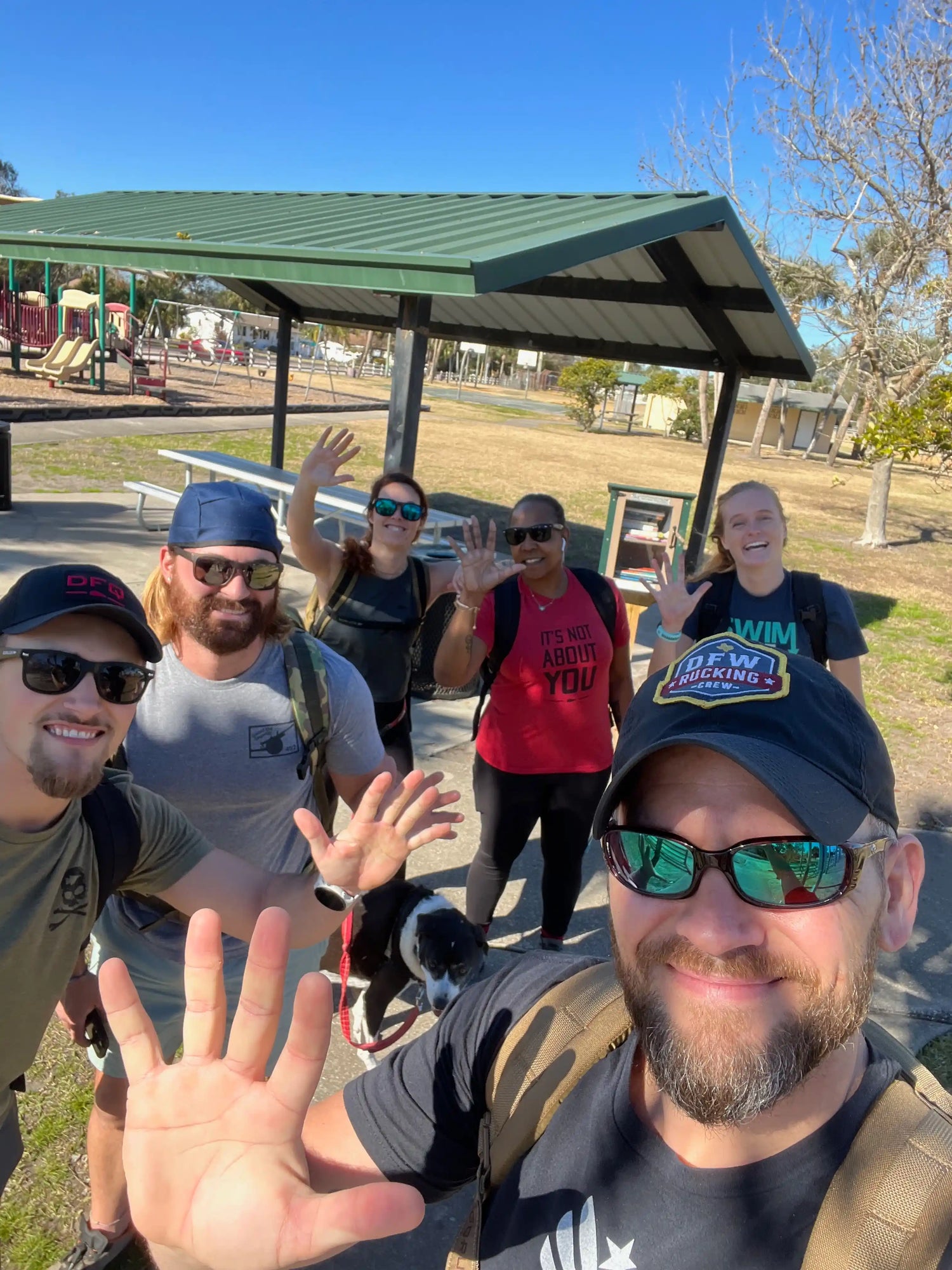 Group of people in GORUCK gear waving at park playground, sunny day, community rucking event