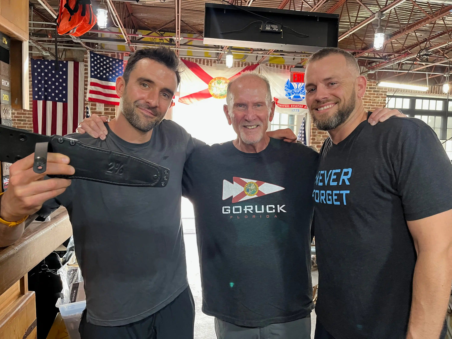 Three men in GORUCK and Never Forget shirts smiling indoors with American and Florida flags behind them