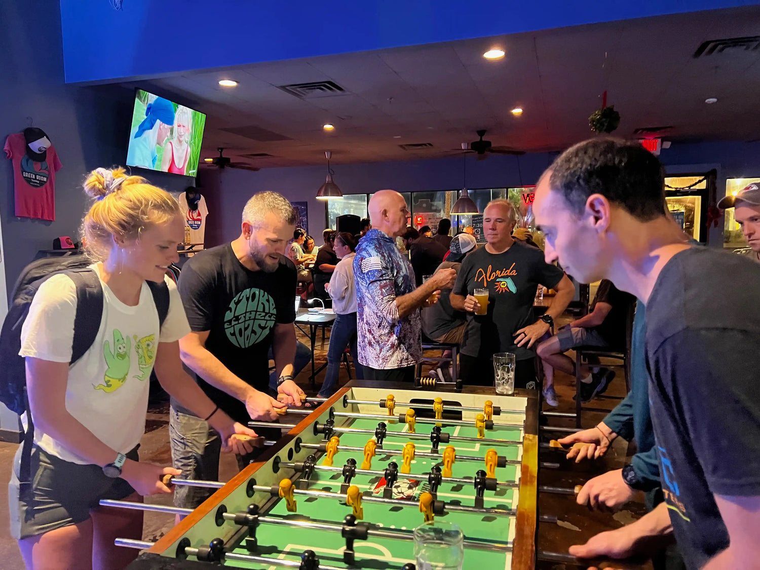Group of adults playing foosball in a lively bar with GORUCK rucking gear and community vibe