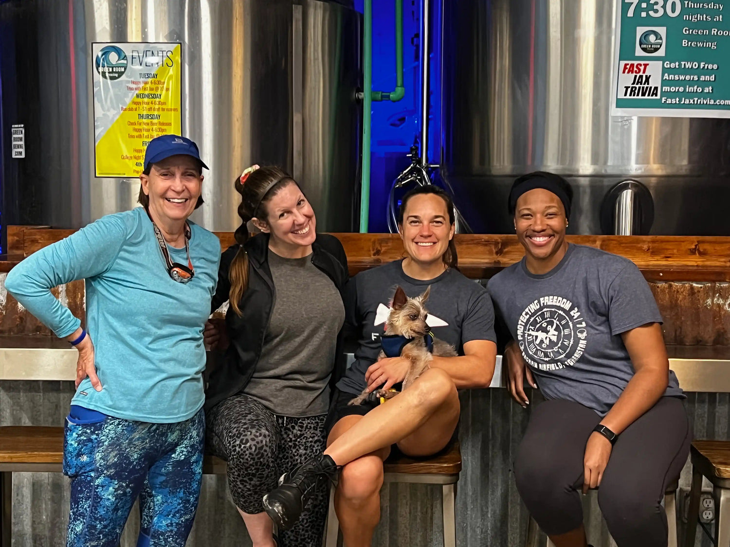 Four women with a small dog in a brewery, wearing athletic GORUCK gear, smiling together.