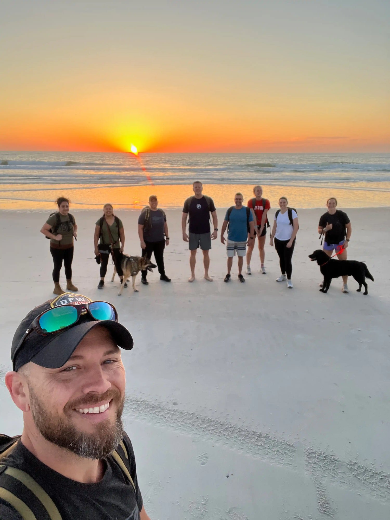 Group of eight people and two dogs on a beach at sunrise, ready for rucking with backpacks