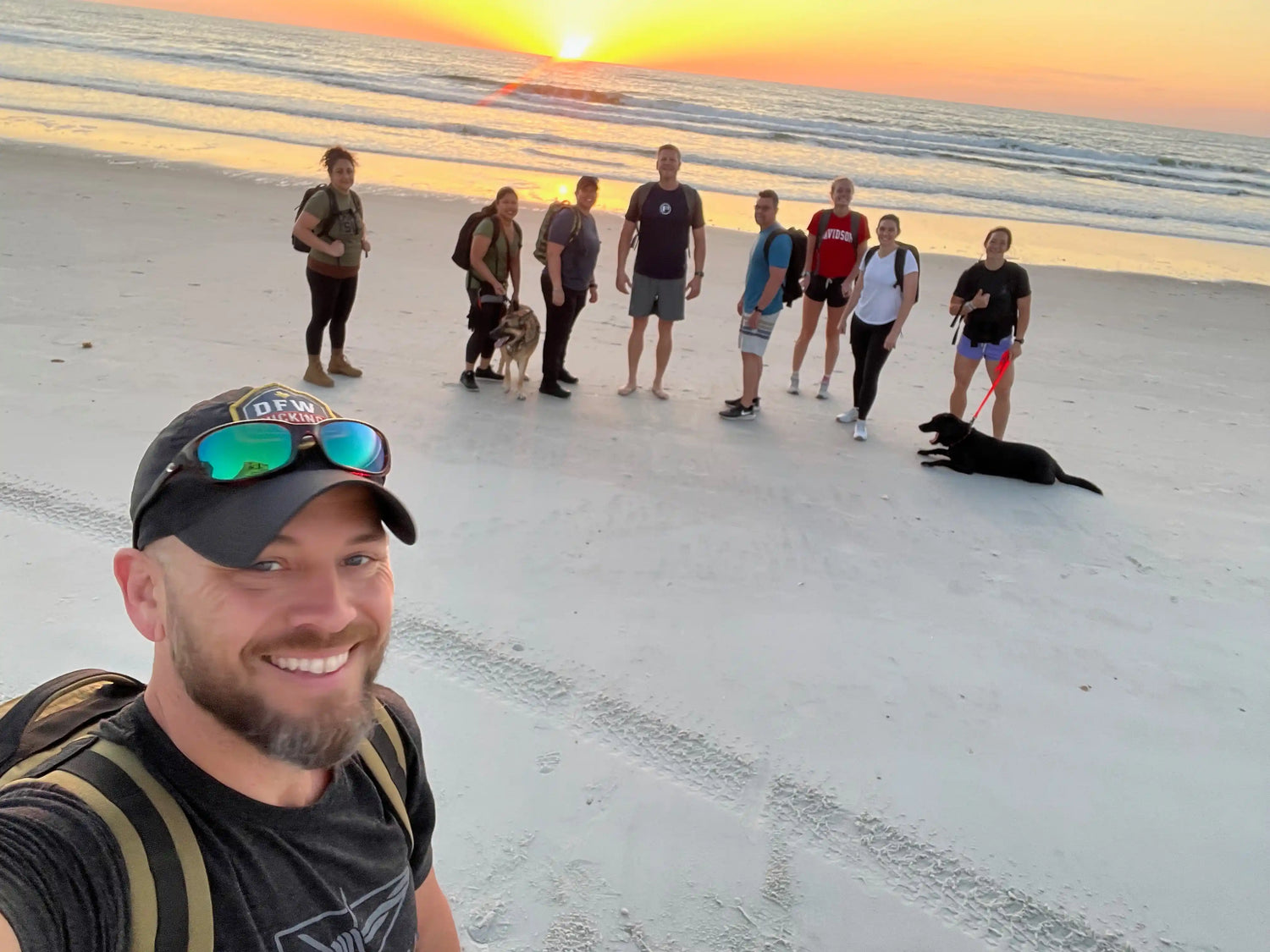 Group rucking on beach at sunrise with GORUCK gear, two dogs, and backpacks visible.
