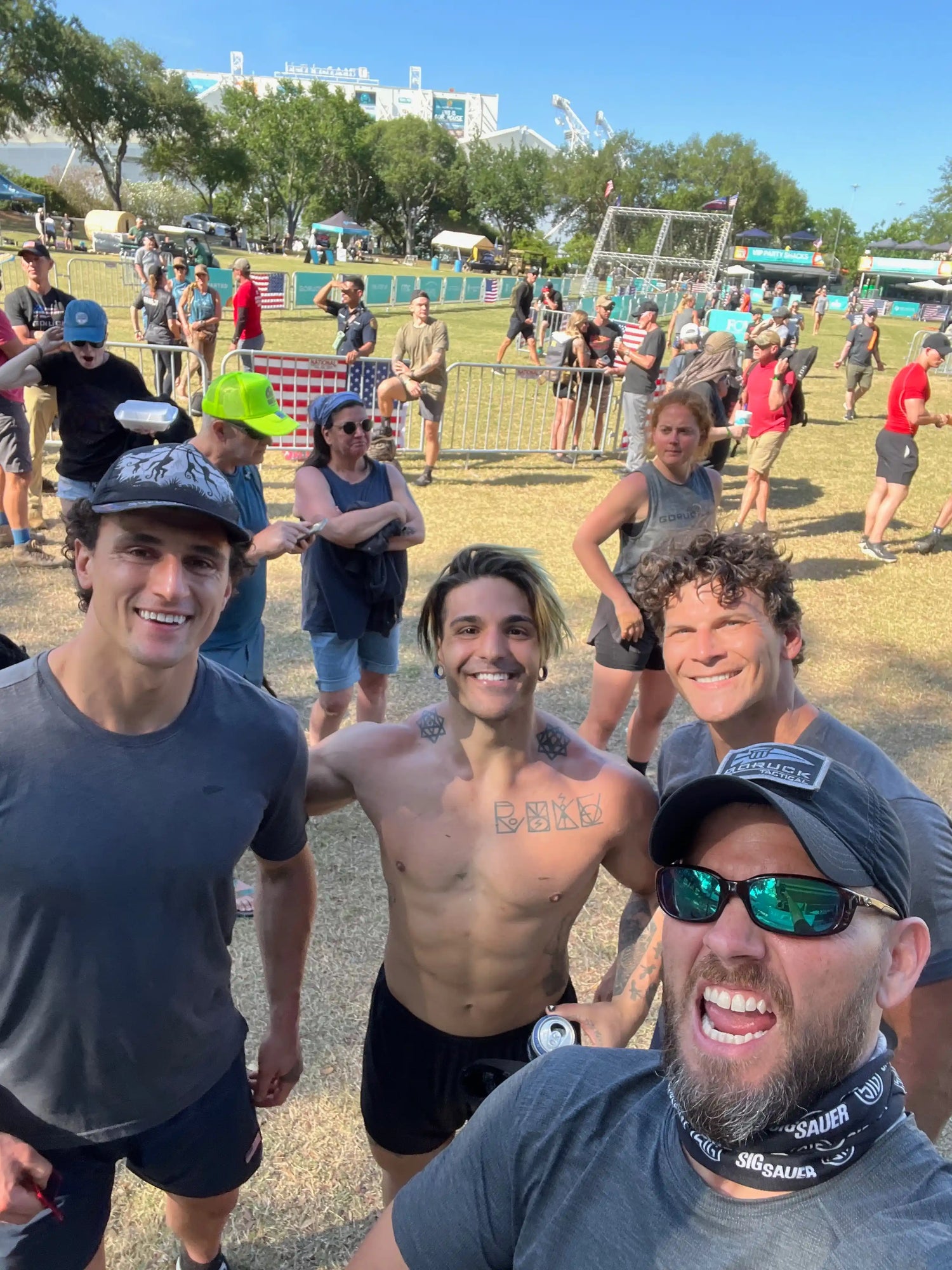 Four men smiling at outdoor event with fitness and competition setups, sunny day