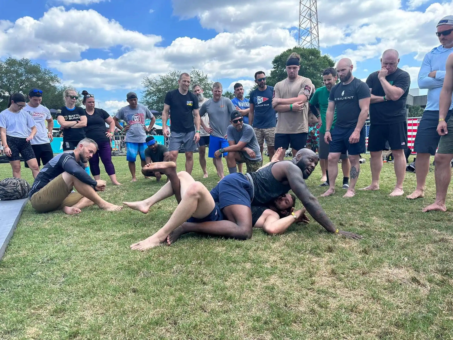 Outdoor grappling training with two men practicing wrestling on grass surrounded by attentive group