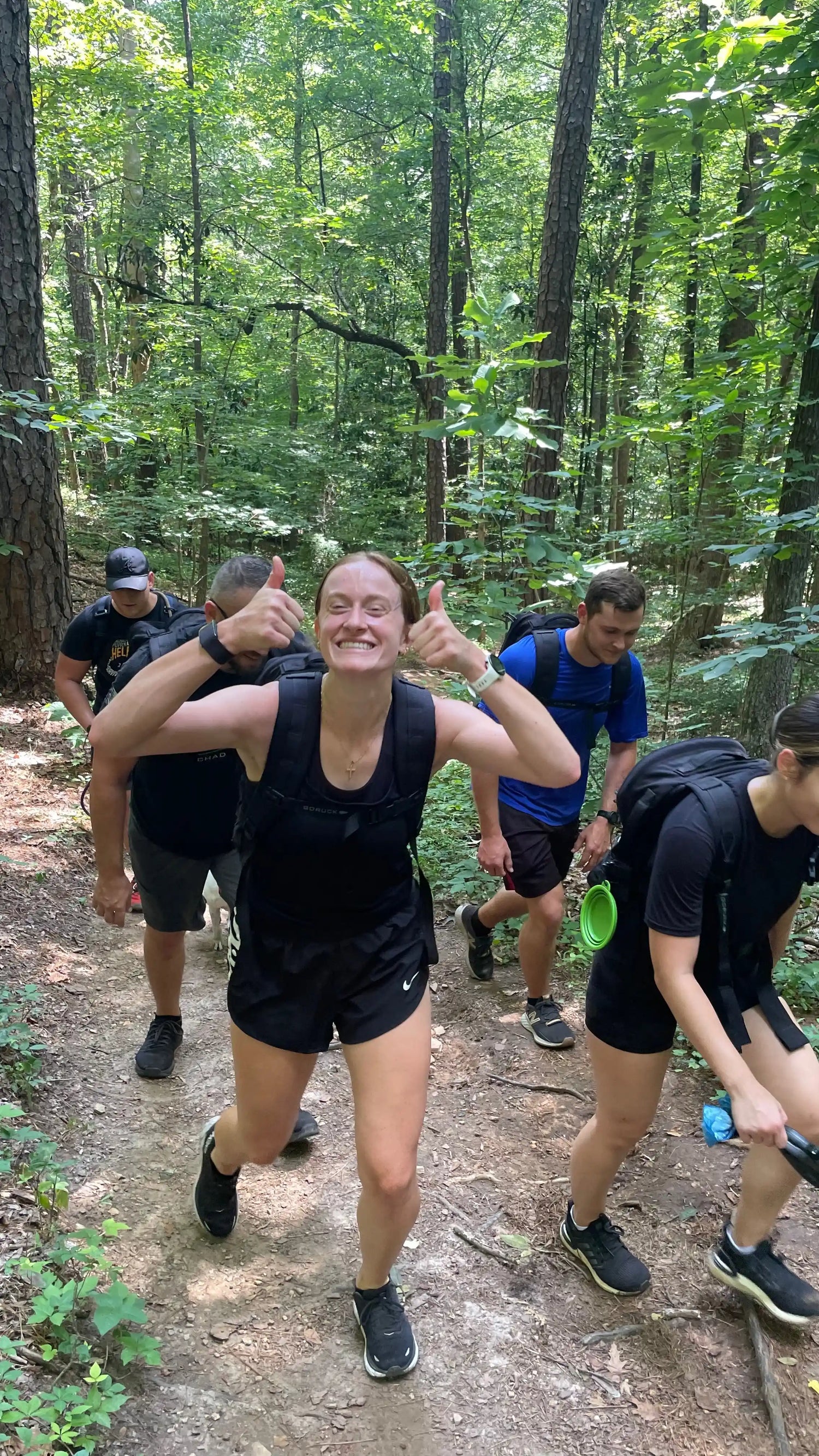 Group rucking with GORUCK backpacks on forest trail, smiling woman gives thumbs up